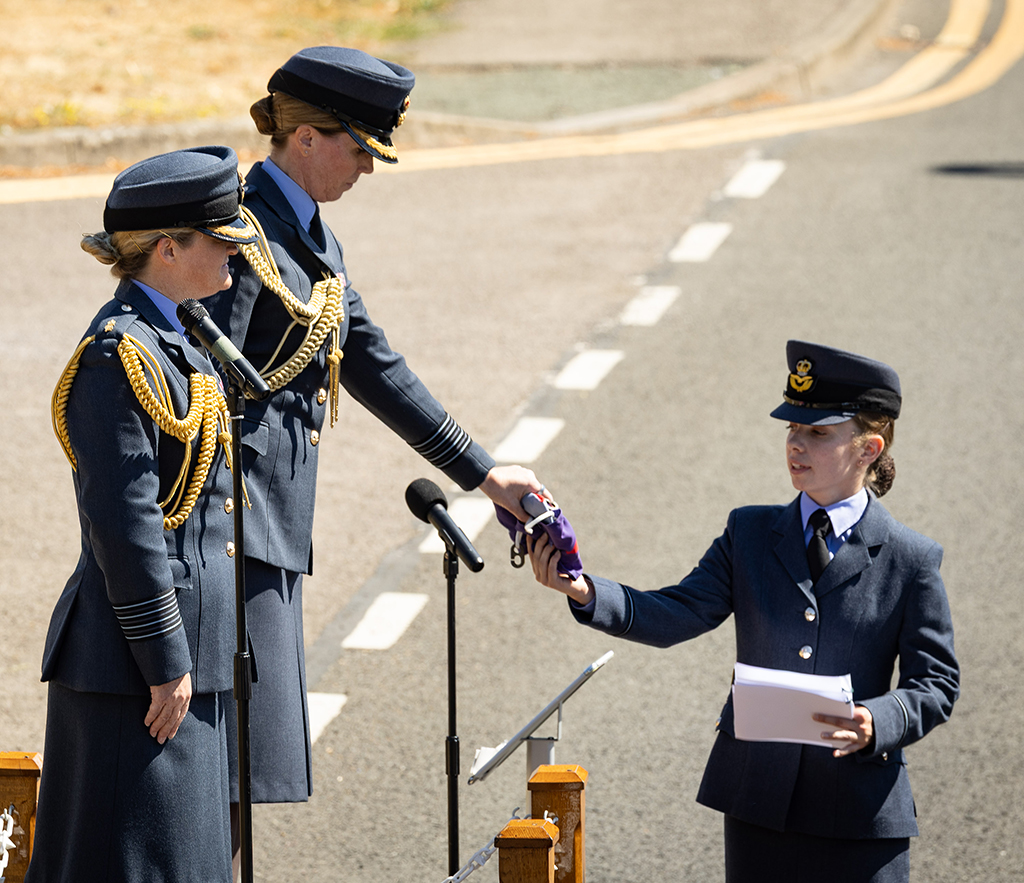 A close up view of Group Captain Louise Henton and Group Captain Kate Macartney, who are both standing on a dais in front of gathered personnel and invited guests. Group Captain Henton is ceremoniously relinquishing the Station Commander's pendant to an officiator.